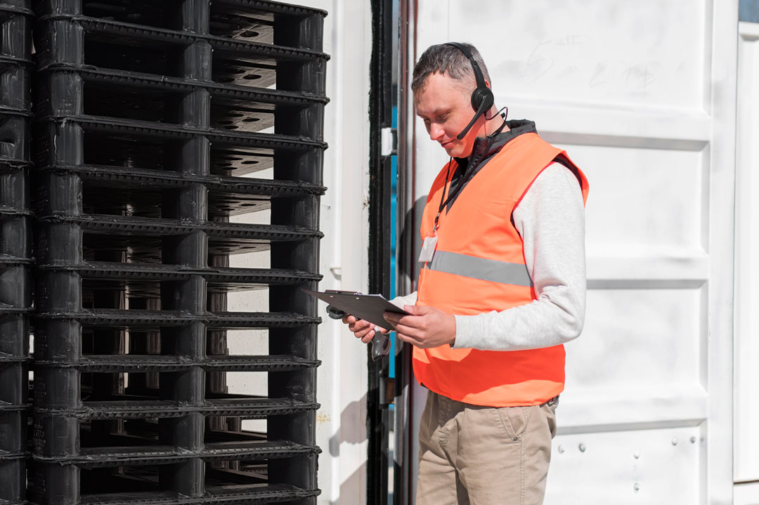 Medium shot of Man in orange vest checking in a shipment of pallets standing outside open freight truck